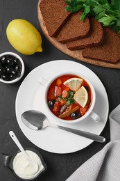 Photo of Meat solyanka soup with sausages, olives and vegetables served on dark grey textured table, flat lay