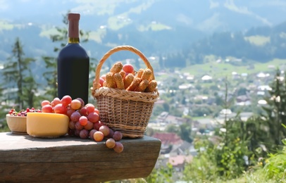 Photo of Bottle of red wine with ripe juicy grapes and other food for picnic on bench against mountain landscape