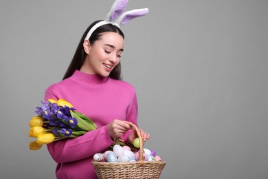 Happy woman in bunny ears headband holding wicker basket with painted Easter eggs and bouquet of flowers on grey background. Space for text