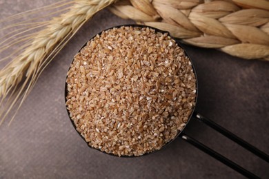 Photo of Dry wheat groats in scoop and spikelet on brown textured table, top view