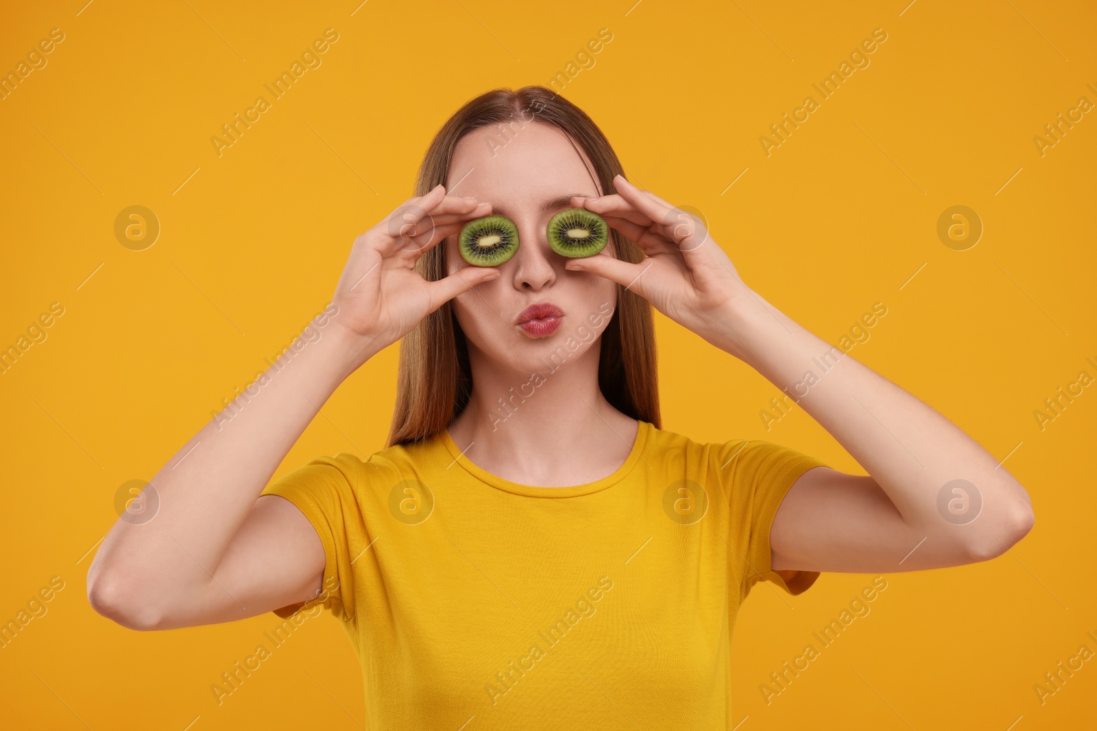 Photo of Young woman holding halves of kiwi near her eyes on yellow background