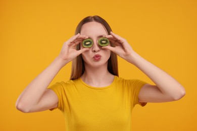 Young woman holding halves of kiwi near her eyes on yellow background