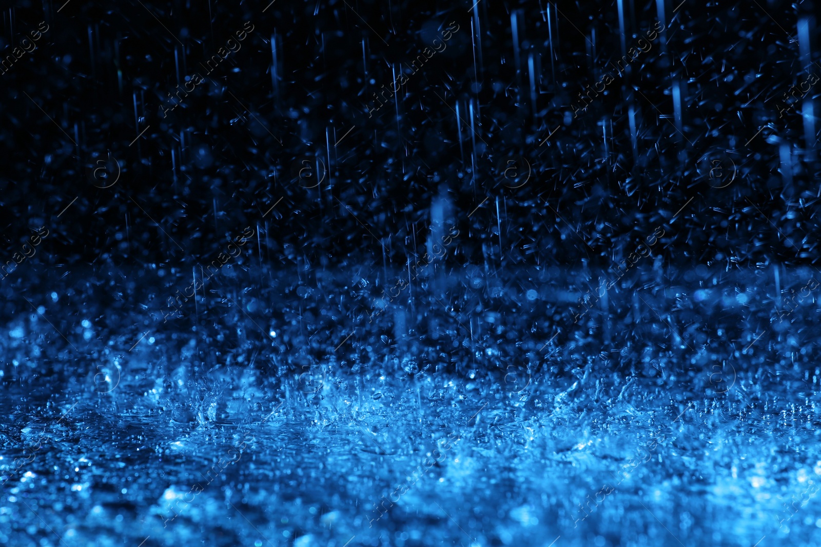 Photo of Heavy rain falling down on ground against dark background, toned in blue