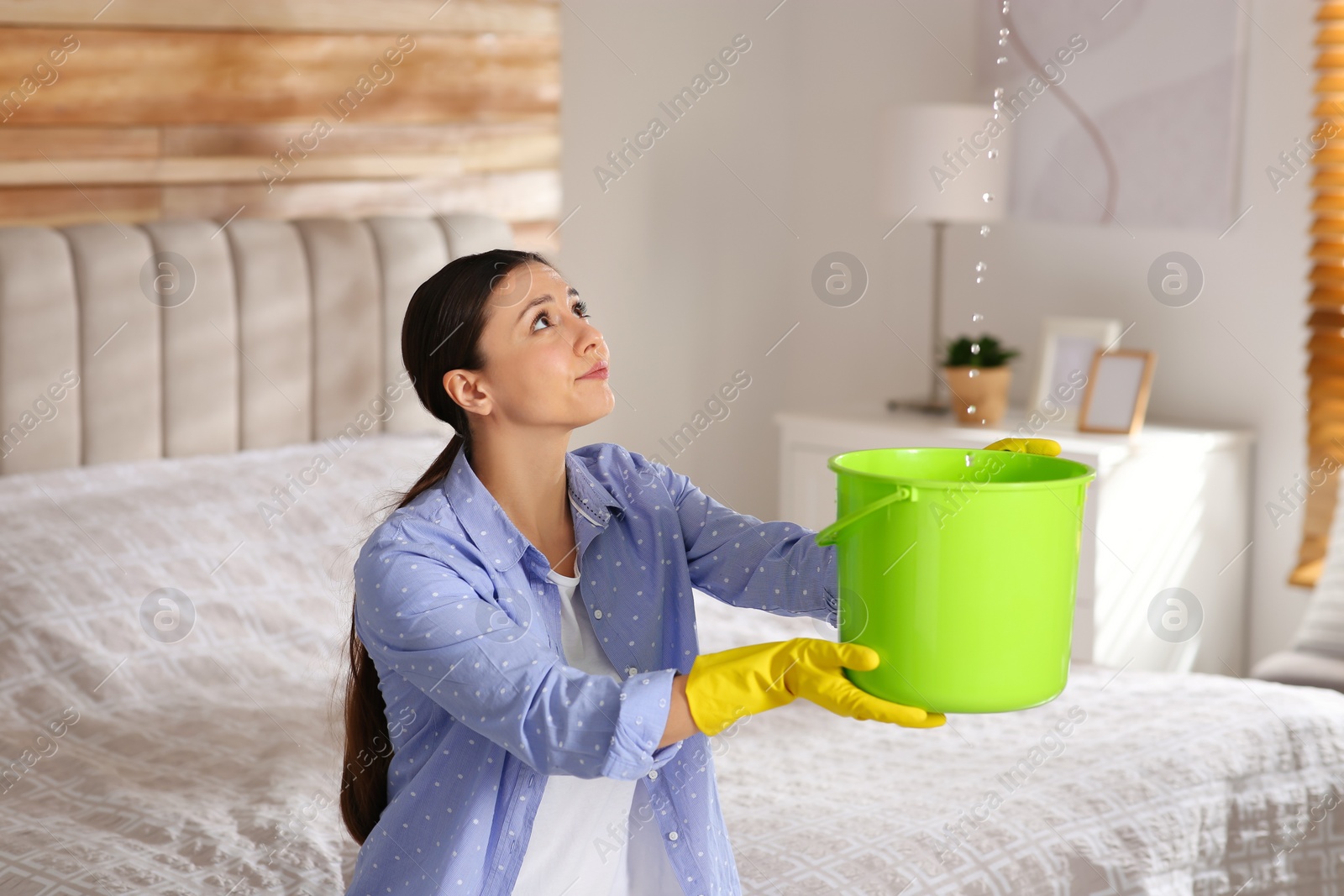 Photo of Young woman collecting leaking water from ceiling in bedroom. Time to call roof repair service