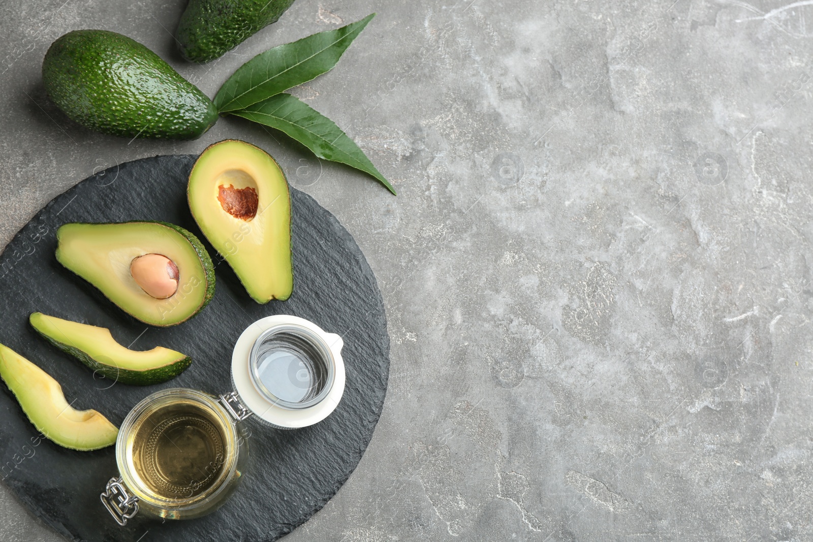 Photo of Board with jar of natural oil and avocados on grey stone background, flat lay. Space for text