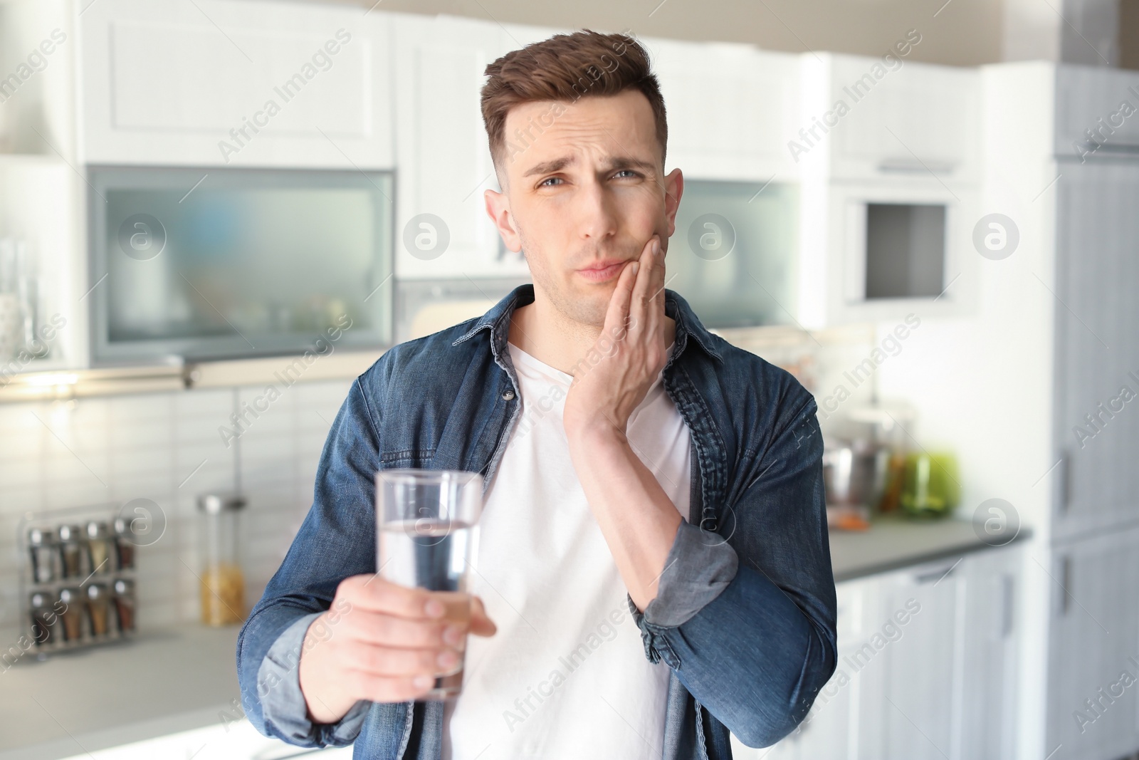 Photo of Young man with sensitive teeth and glass of cold water in kitchen