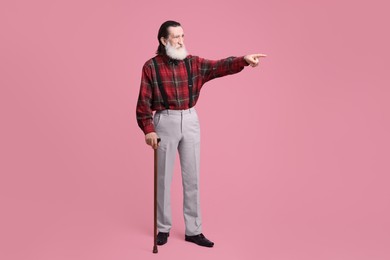 Senior man with walking cane pointing at something on pink background