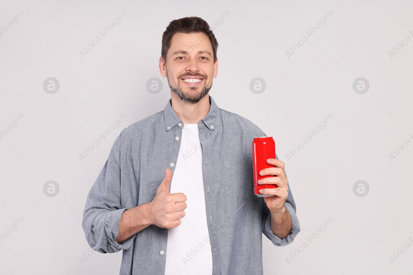 Photo of Happy man holding red tin can with beverage and showing thumb up on light grey background