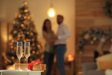 Image of Glasses with champagne and Christmas gift on table and couple in room, bokeh effect. Space for text