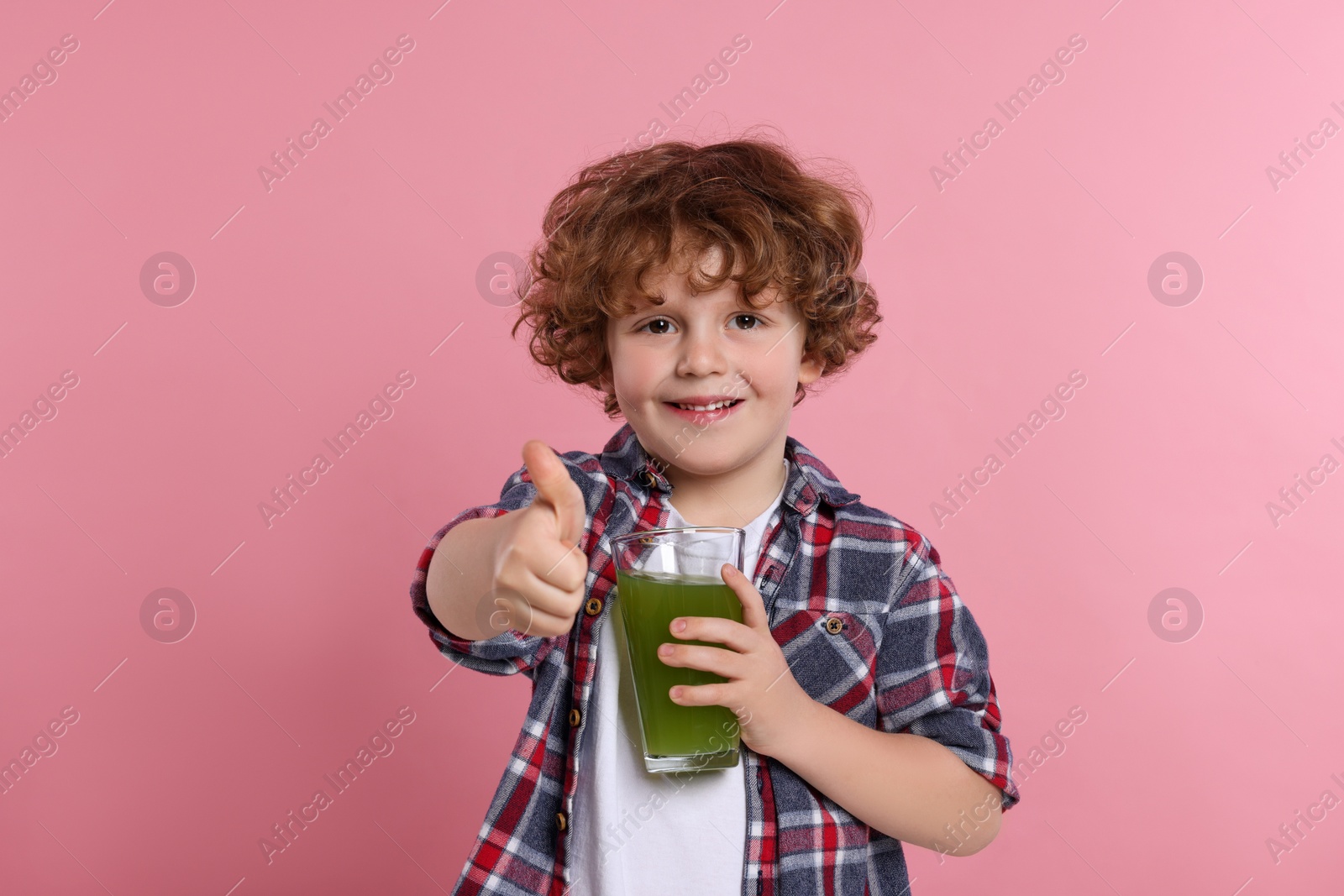 Photo of Cute little boy with glass of fresh juice showing thumb up on pink background
