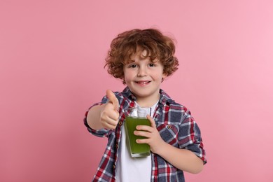 Cute little boy with glass of fresh juice showing thumb up on pink background