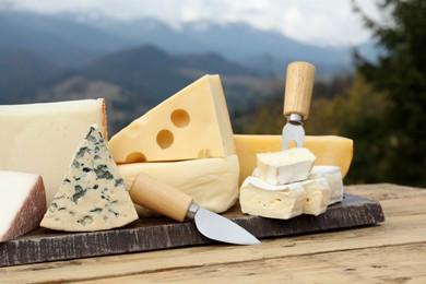 Different types of delicious cheeses on wooden table against mountain landscape