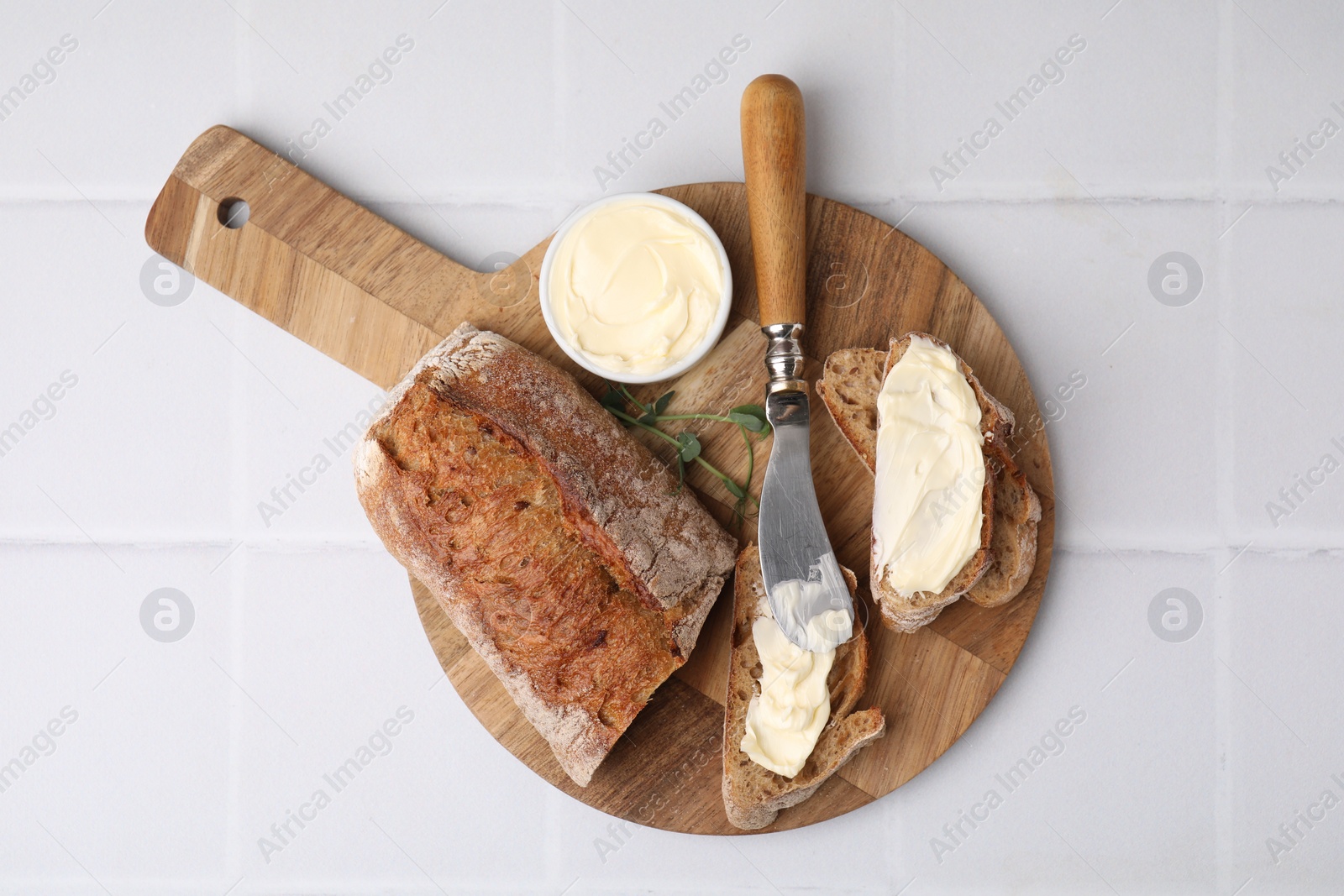 Photo of Tasty bread with butter and knife on white tiled table, top view