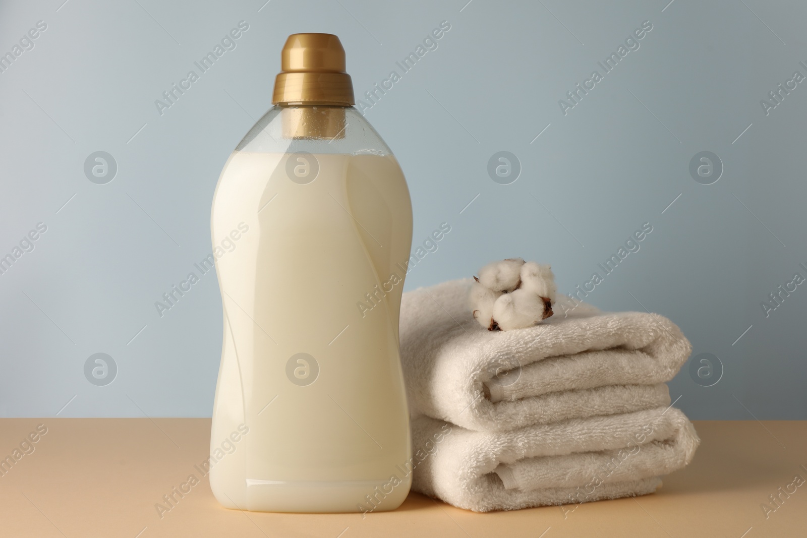 Photo of Bottle of fabric softener, fluffy cotton flower and stacked clean towels on pale yellow table against light grey background