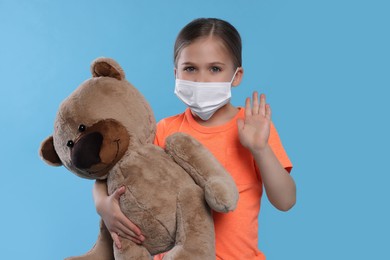 Photo of Cute girl with medical mask and teddy bear on light blue background