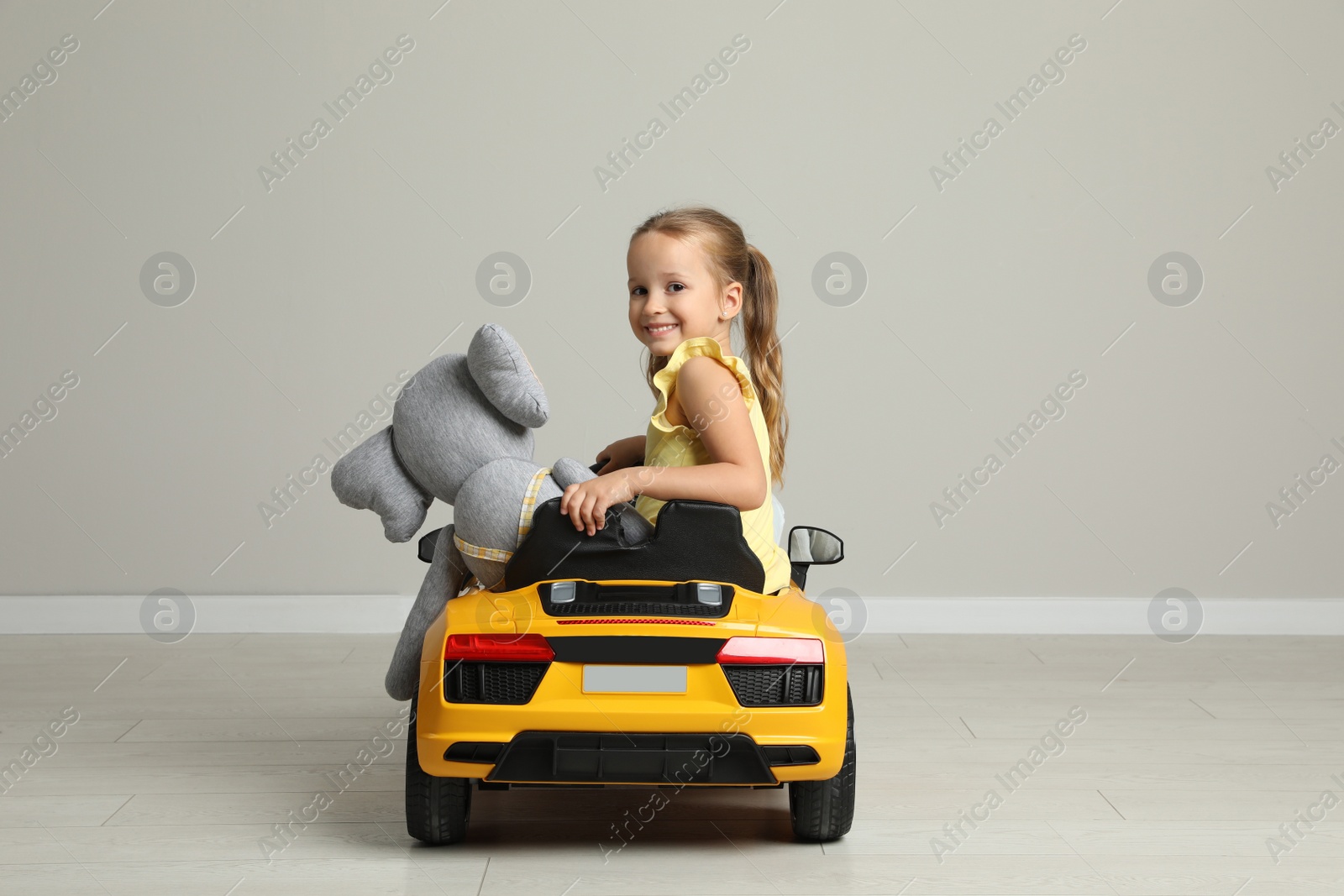 Photo of Cute little girl with toy elephant driving children's car near grey wall indoors