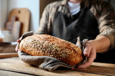 Photo of Man holding loaf of fresh bread at wooden table indoors, closeup