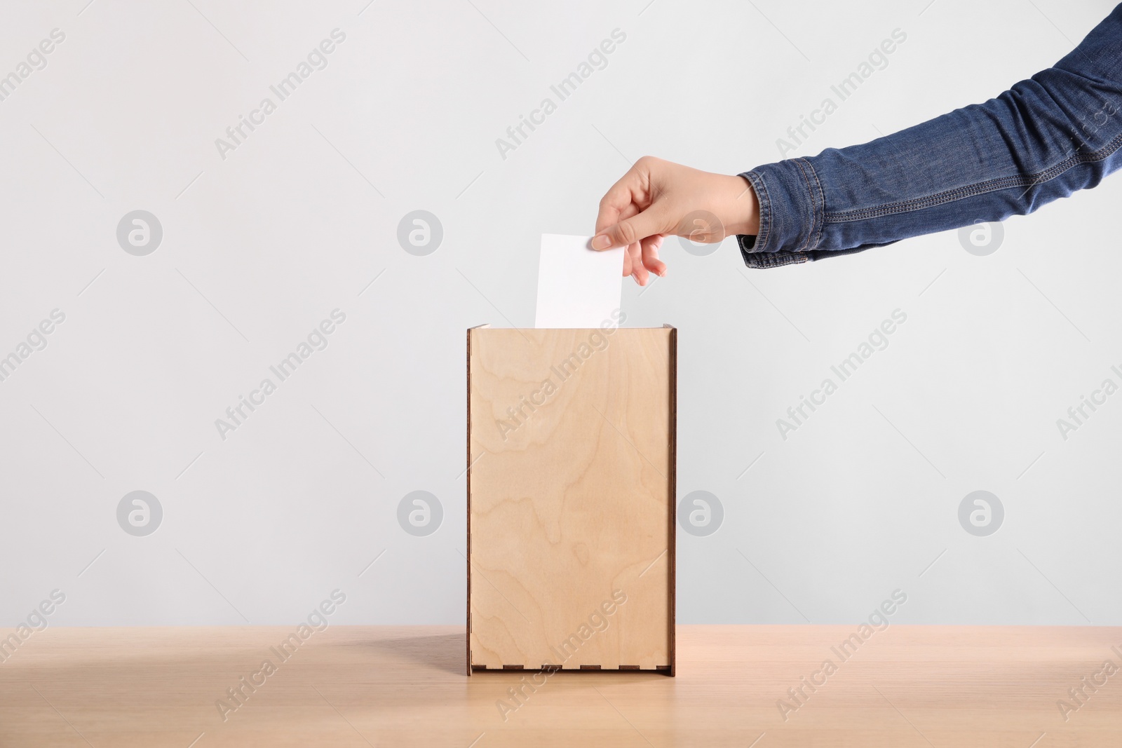 Photo of Woman putting her vote into ballot box on wooden table against light grey background, closeup