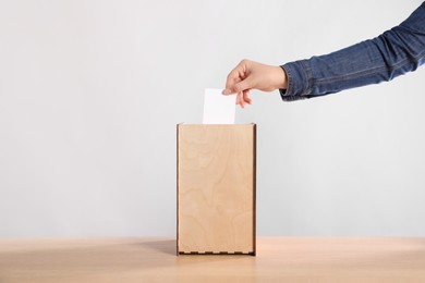 Woman putting her vote into ballot box on wooden table against light grey background, closeup
