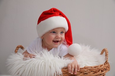 Photo of Cute baby wearing Santa hat in wicker basket on light grey background. Christmas celebration