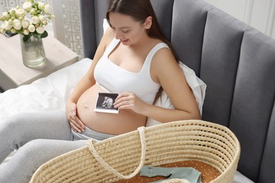 Photo of Beautiful pregnant woman with ultrasound picture of baby and basket on bed indoors