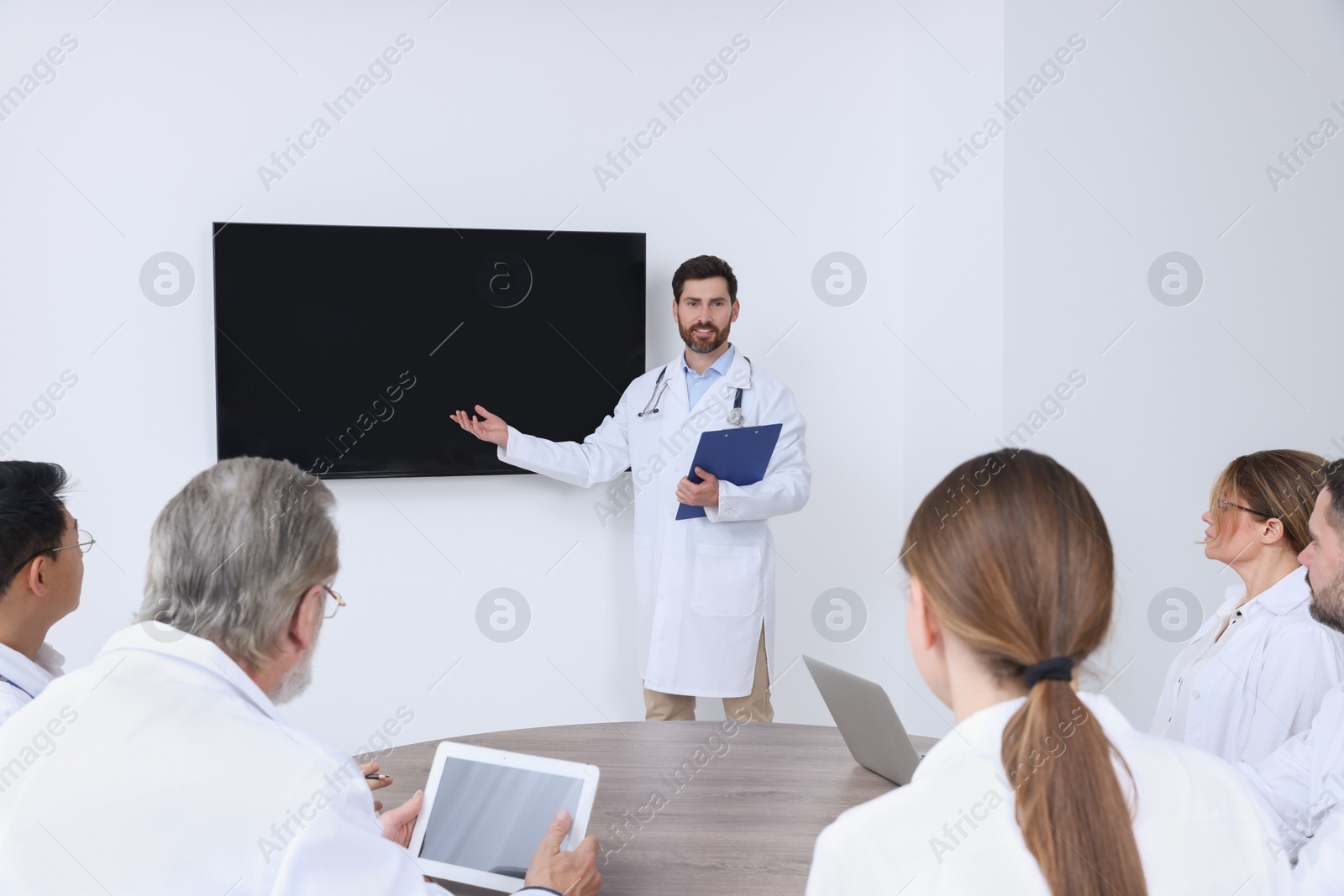 Photo of Team of doctors listening to speaker report near tv screen in meeting room. Medical conference