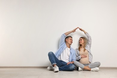 Photo of Pregnant woman with her husband forming roof with their hands while sitting on floor indoors. Space for text