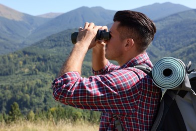 Tourist with backpack and sleeping pad looking through binoculars in mountains