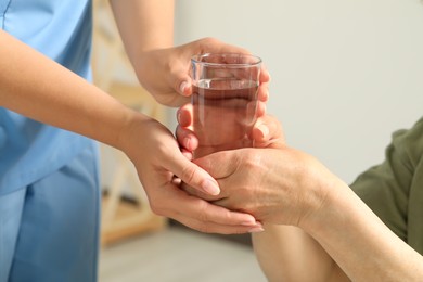 Caretaker giving glass of water to elderly woman indoors, closeup