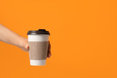 Photo of Woman holding takeaway paper coffee cup with cardboard sleeve on orange background, closeup. Space for text