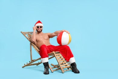 Muscular young man in Santa hat with ball, sunglasses and deck chair on light blue background, space for text