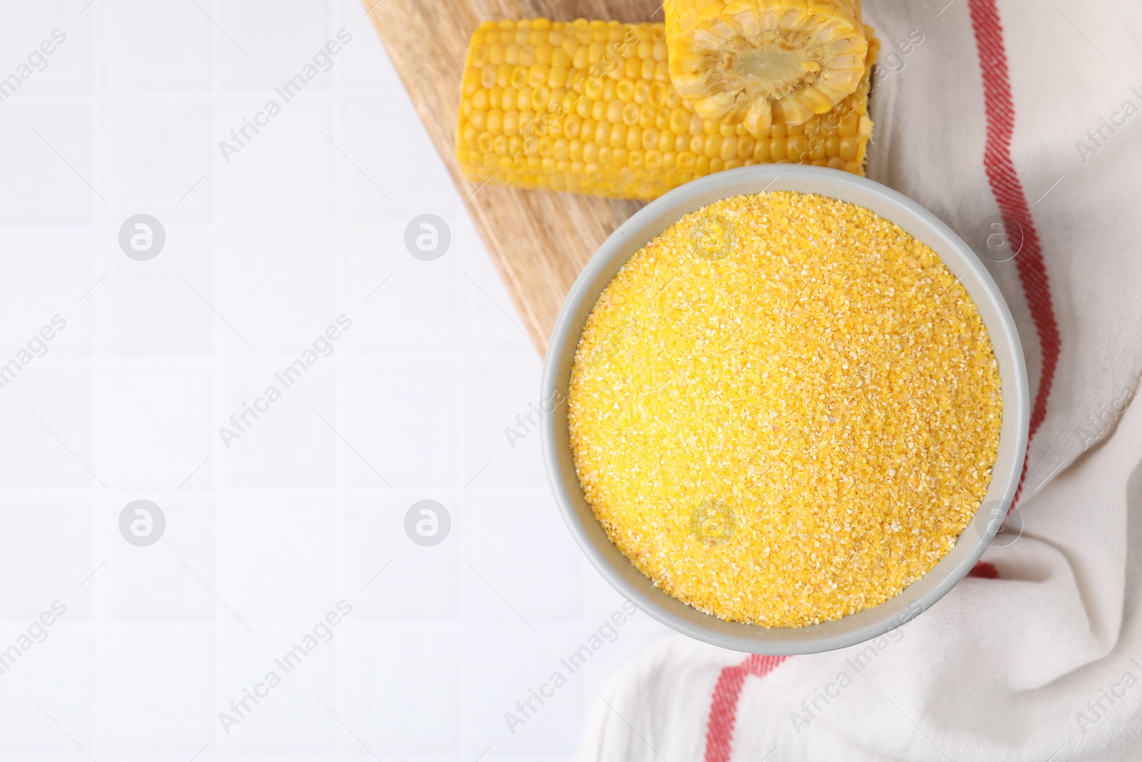 Photo of Raw cornmeal in bowl and corn cobs on white tiled table, top view. Space for text