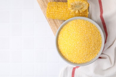 Raw cornmeal in bowl and corn cobs on white tiled table, top view. Space for text