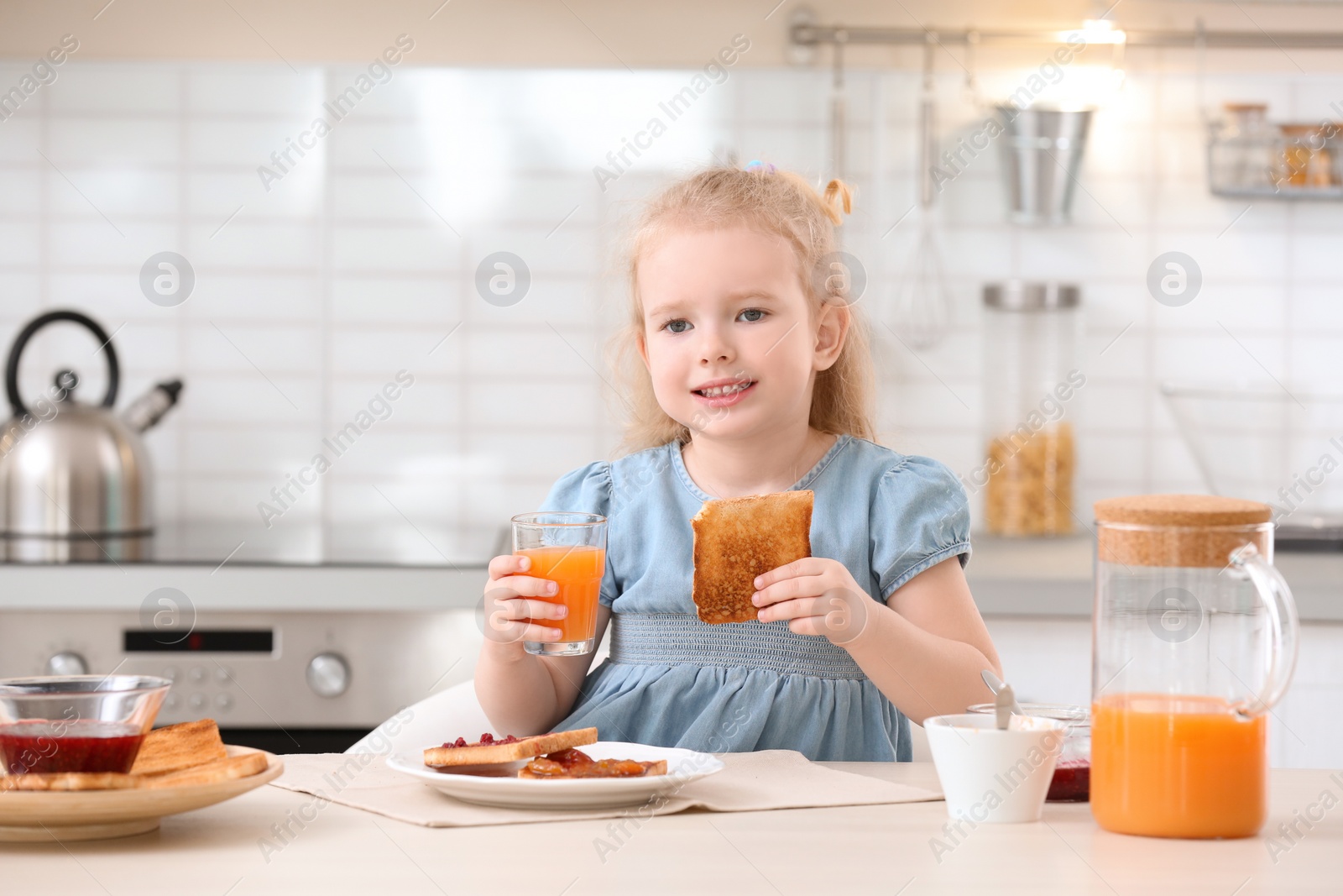 Photo of Adorable little girl eating tasty toasted bread with jam at table in kitchen