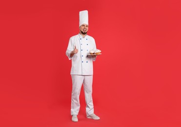 Photo of Happy professional confectioner in uniform holding delicious cupcakes and showing thumbs up on red background