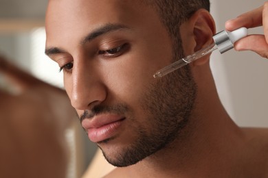 Photo of Handsome man applying cosmetic serum onto face indoors, closeup