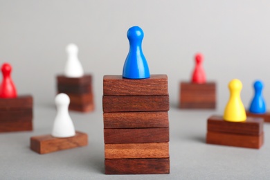 Photo of Board game piece on wooden blocks dominating other figures against grey background. Victory concept