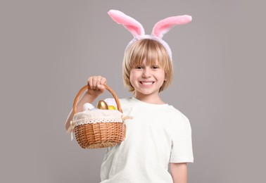 Photo of Happy boy in bunny ears headband holding wicker basket with painted Easter eggs on grey background