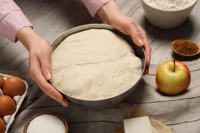 Photo of Woman holding bowl with fresh yeast dough and ingredients for cake on wooden table, closeup