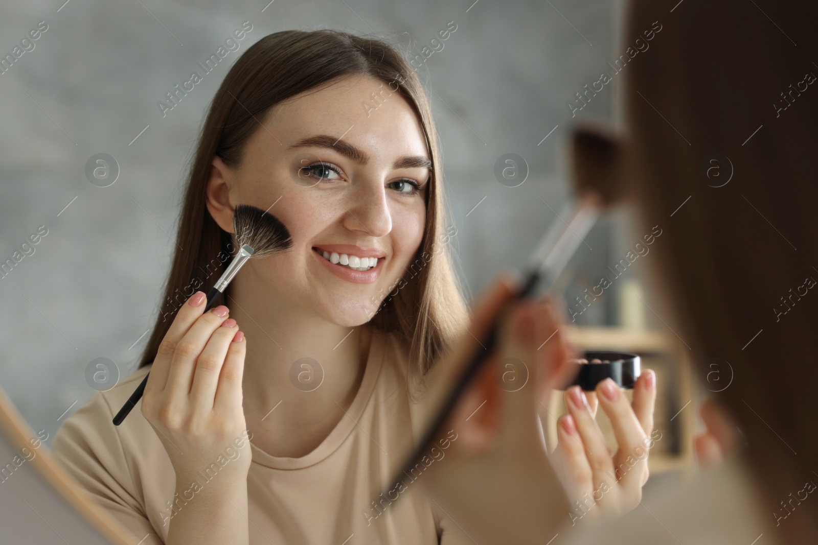 Photo of Smiling woman with freckles applying makeup near mirror indoors