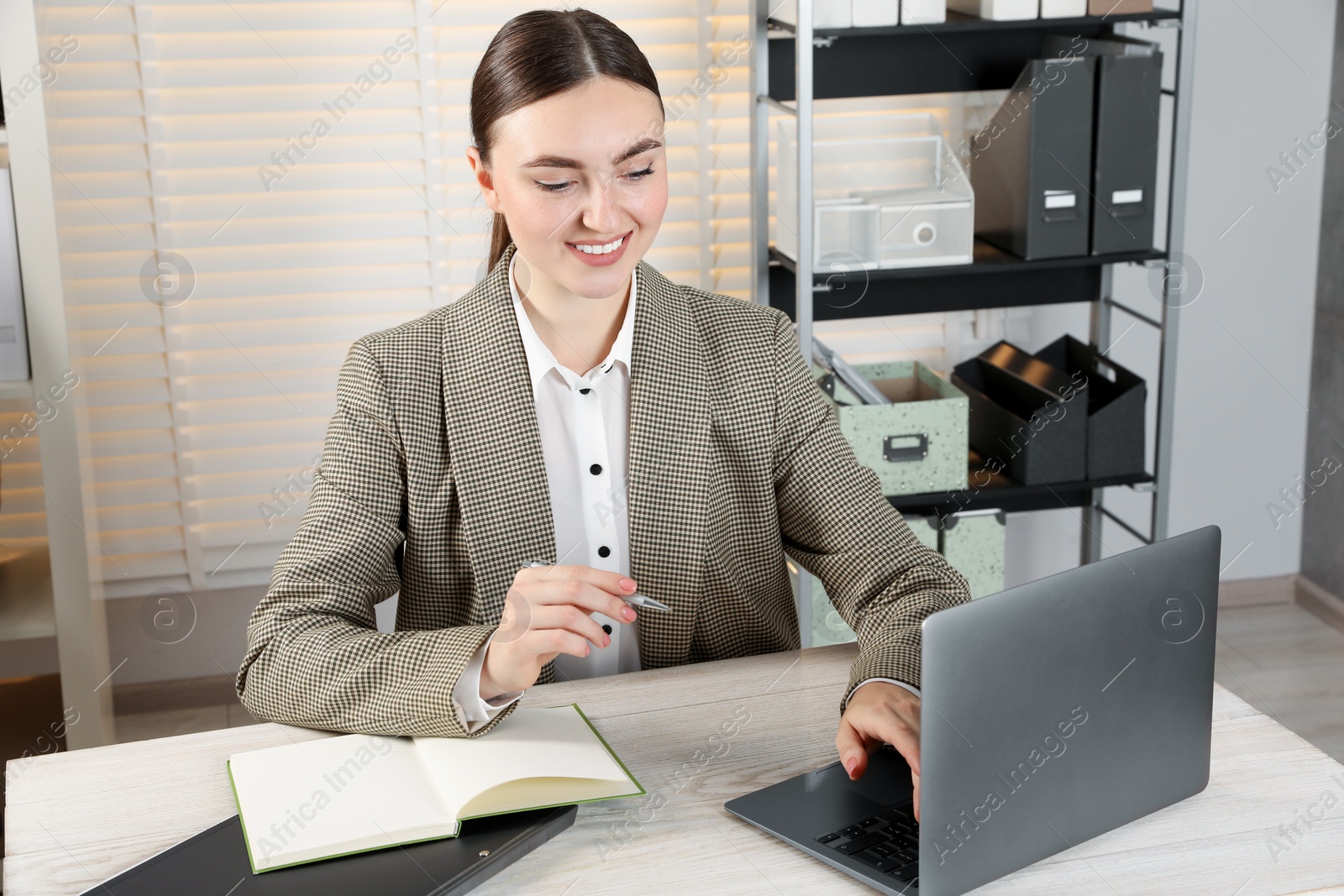 Photo of Happy woman taking notes while using laptop at light wooden table in office