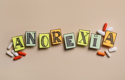 Photo of Word Anorexia made of papers with letters near pills on beige background, flat lay