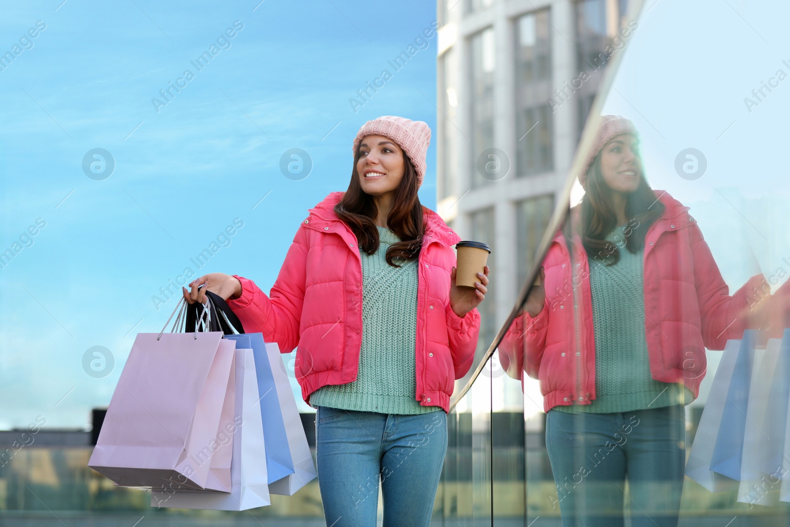 Photo of Beautiful young woman with cup of coffee and shopping bags near building