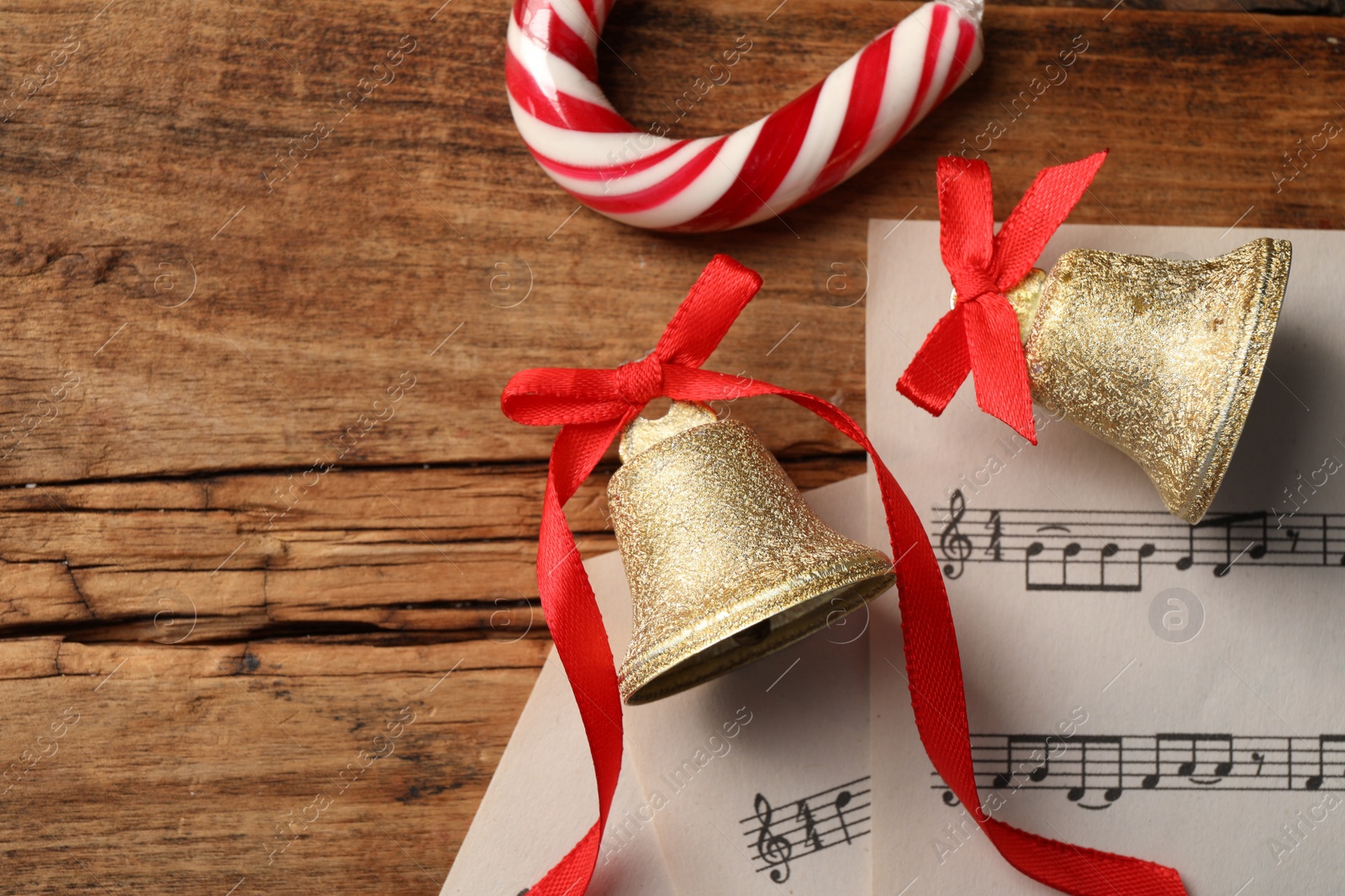 Photo of Golden shiny bells with red bows, music sheets and candy cane on wooden table, flat lay. Space for text. Christmas decoration