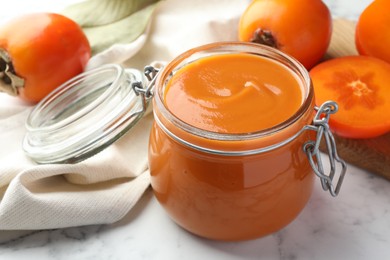 Photo of Delicious persimmon jam and fresh fruits on white marble table, closeup