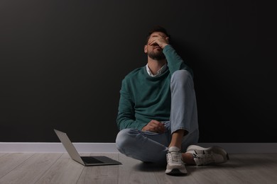 Photo of Upset man sitting on floor near laptop against black wall. Space for text