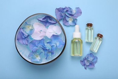 Spa composition. Aromatic water in bowl, flowers and bottles of essential oil on light blue background, flat lay