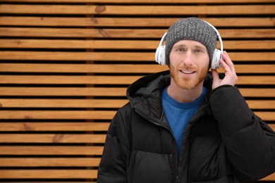 Photo of Young man listening to music with headphones against wooden wall. Space for text