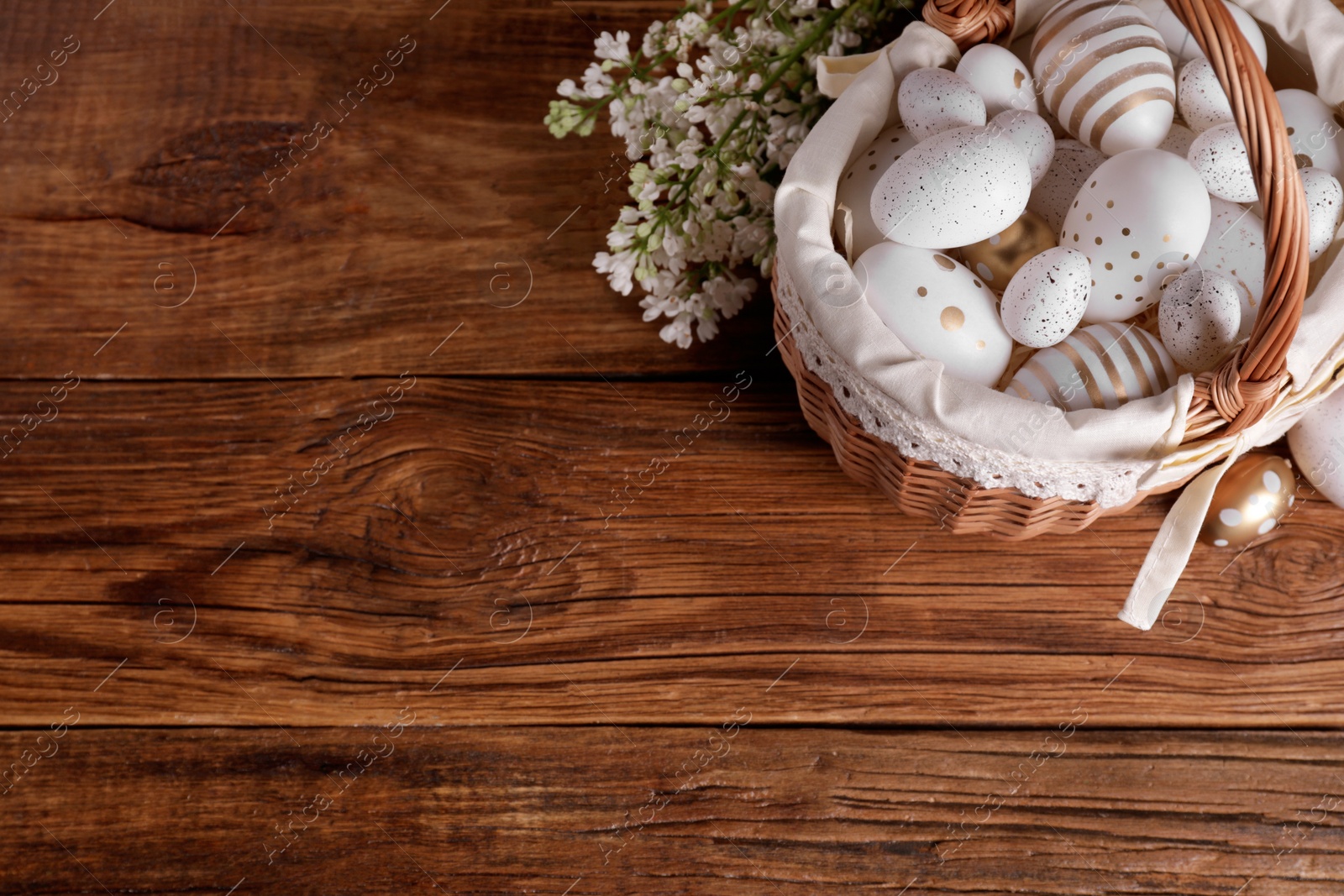 Photo of Wicker basket with festively decorated Easter eggs and white lilac flowers on wooden table, flat lay. Space for text