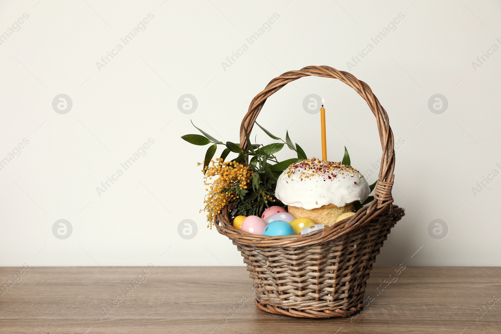 Photo of Traditional Easter cake with burning candle, dyed eggs and flowers in basket on table against white background. Space for text